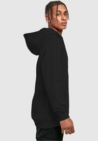 Black hoodie with a kangaroo pocket, featuring a hood and ribbed cuffs. Smooth texture and relaxed fit, shown on a model in profile.