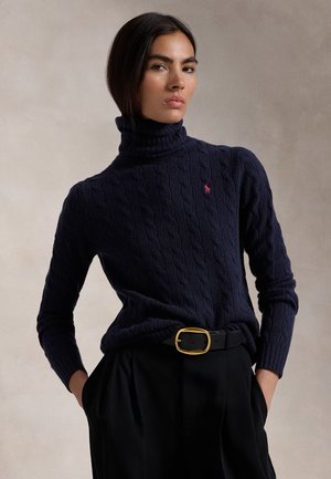 Femme portant un pull à col roulé en maille torsadée bleu marine avec un logo rouge, une ceinture noire avec une boucle dorée et un pantalon noir, mains dans les poches.