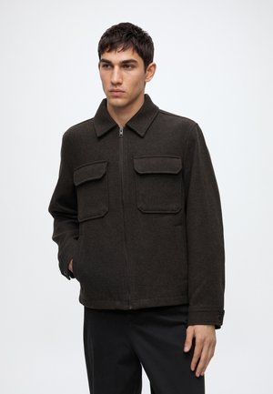 Jeune homme portant une veste zippée marron foncé avec deux poches poitrine et un pantalon noir, debout devant un fond clair uni.
