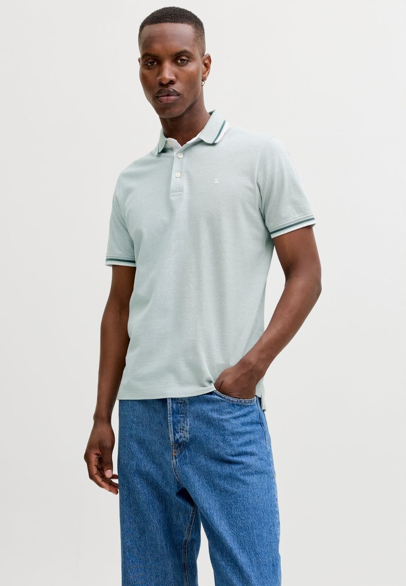 Homme aux cheveux courts portant un polo vert clair et un jean bleu, une main dans la poche, debout devant un fond uni.
