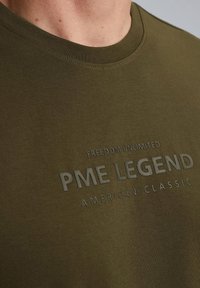 Maslinasto zelena pamučna majica s okruglim izrezom. Sadrži podignut, u reljefu otisnut tekst: "NEOGRANIČENA SLOBODA PME LEGEND AMERICAN CLASSIC" u svijetlosivoj boji.