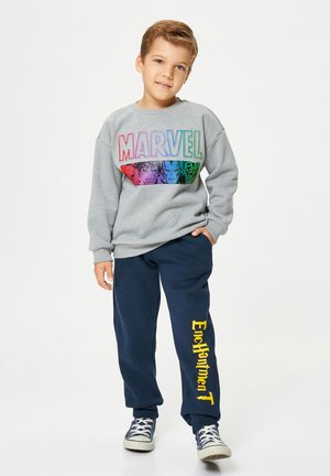 Junge steht mit einer Hand in der Tasche, trägt einen grauen Marvel-Sweatshirt und marineblaue Jogginghosen mit gelbem "Enchantment"-Text sowie marineblauen Sneakers.