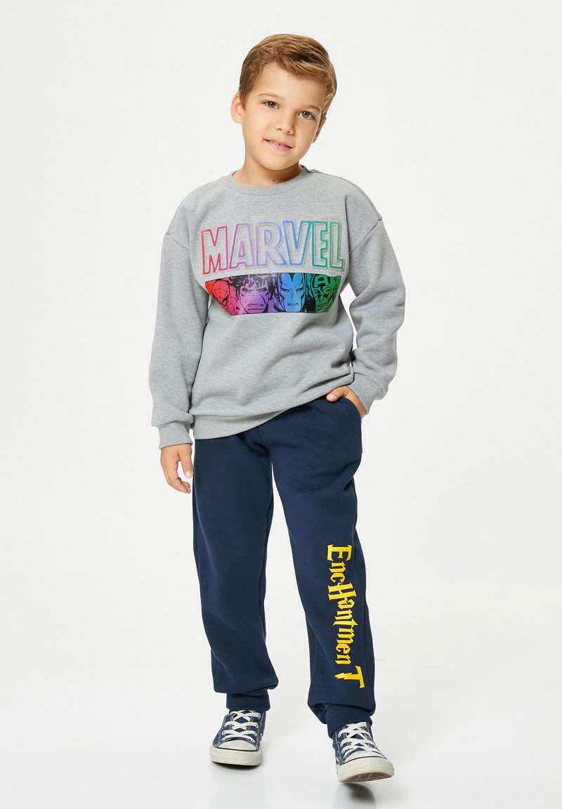 Chico de pie con una mano en el bolsillo, vistiendo una sudadera gris de Marvel y un pantalón de chándal azul marino con el texto amarillo "Enchantment", calzado con zapatillas de deporte azul marino.