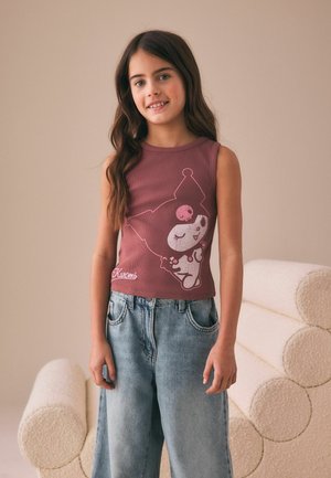 Ung pige med langt brunt hår iført en ærmeløs lilla top med tegneseriefigur og løse lyseblå jeans, står indendørs.