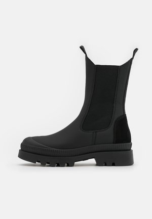 Schwarzer Stiefel bis zur Mitte der Wade mit Profilsohle, elastischen Seitenteilen, Zuglaschen, abgerundetem Zehenbereich und dicker, strukturierter Gummisohle vor weißem Hintergrund.