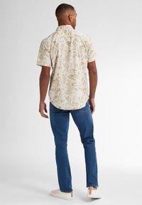 Camisa de manga corta de color claro con un patrón de hojas, combinada con jeans azules y zapatillas blancas, vista desde la parte de atrás.