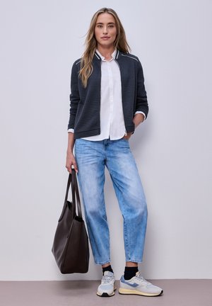 Frau trägt schwarze Bomberjacke, weißes Hemd, hellblaue Jeans, weiße Sneaker, hält große dunkelbraune Tote-Bag, steht vor einfachem Hintergrund.