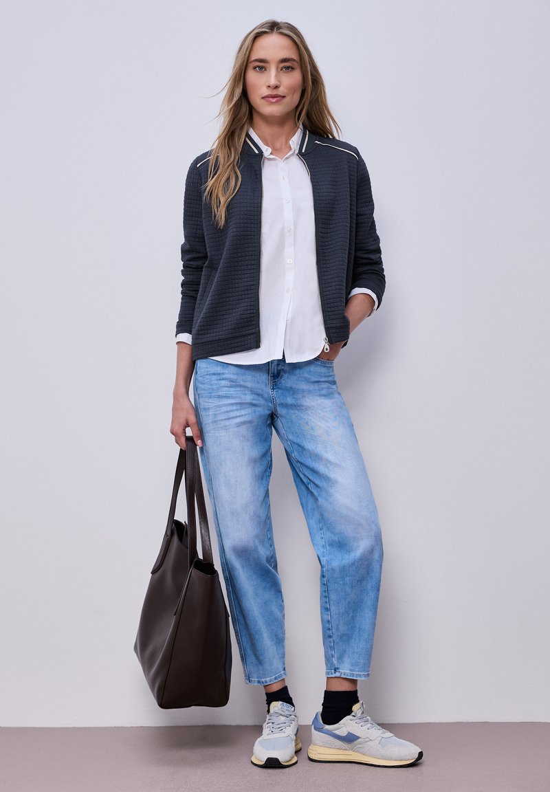 Femme portant une veste bomber noire, une chemise blanche, un jean bleu clair, des baskets blanches, tenant un grand sac fourre-tout marron foncé, debout devant un fond uni.