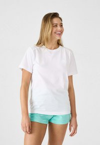 Camiseta de algodón blanca de manga corta y cuello redondo, con un pequeño logo. Combinada con pantalones cortos de color azul verdoso.