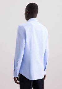 Camicia azzurro chiaro a maniche lunghe realizzata in tessuto morbido, con colletto classico e polsini con bottoni, dotata di un fondo curvo. Nessun motivo visibile.