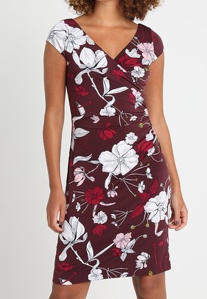 Kvinde iført en burgunderfarvet knælange kjole med hvide, lyserøde og røde blomsterprint og korte ærmer.