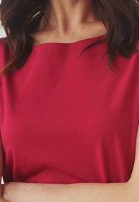 Nainen, jolla on olkapituinen tumma hius, pukeutuneena punaiseen venepääntiepaitaan, kädet ristinä kuvan alareunassa.