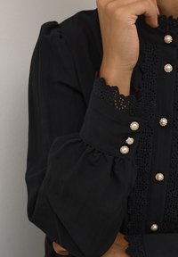Blouse noire avec des manches bouffantes, des accents en dentelle, des boutons en perles et des détails en dentelle complexes le long de l'avant et des poignets.