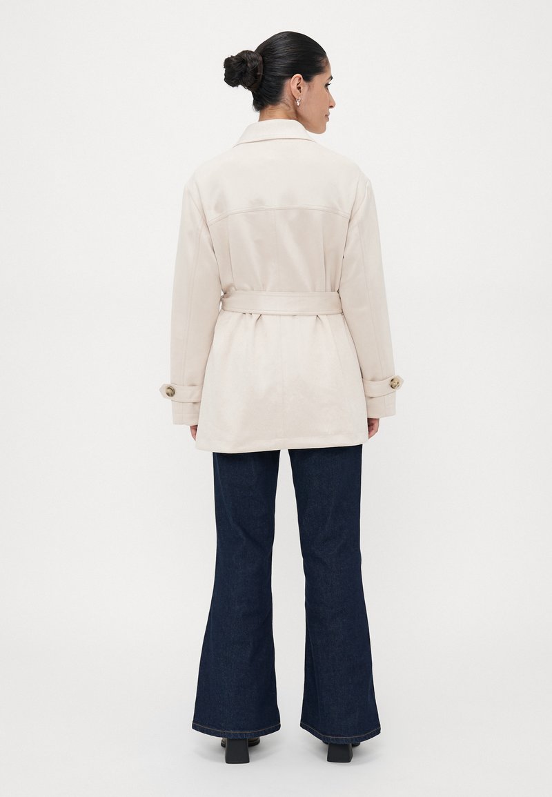 Beige Trenchcoat mit einem taillierten Gürtel, großen Knöpfen an den Ärmeln und einem klassischen Kragen; kombiniert mit dunklen Schlagjeans.