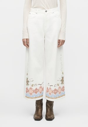 Białe spodnie o szerokich nogawkach z kolorowymi haftowanymi wzorami kwiatowymi i geometrycznymi przy nogawkach, zestawione z brązowymi botkami i długim rękawem.
