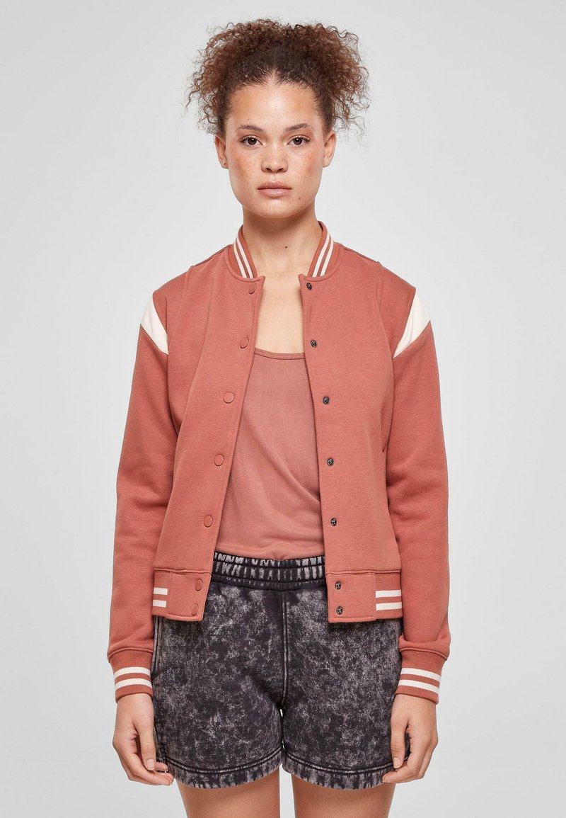 Roostevärvi bomber-tüüpne jakk, valgete õlakaunistustega, trukkidega ja triibuliste soonikkoes mansettidega, kandmas helepluusat ja tumeda mustriga lühikesi pükse.