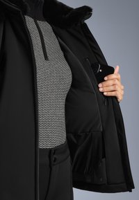 Crna izolirana jakna s krznenim ovratnikom, koja ima džep s patentnim zatvaračem i uzorčanu sivu majicu ispod. Glatka tekstura, puni prednji patent.