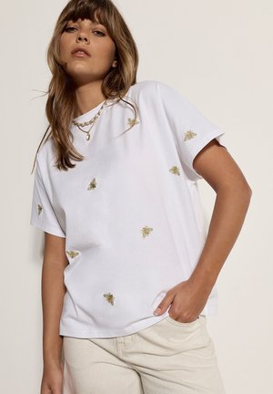 Mujer con camiseta blanca con pequeños motivos de abejas doradas, collares dorados en capas y pantalones beige, posando con la mano en el bolsillo.