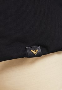Tissu noir avec un motif texturé, présentant une étiquette logo en forme de cœur dorée et noire. Tissu beige lisse visible en dessous.