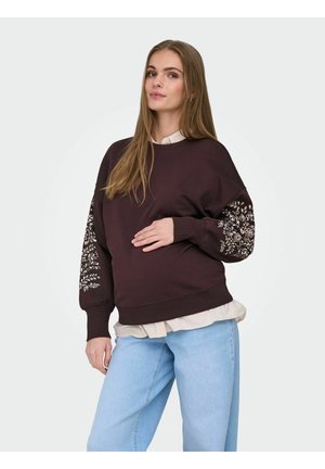 Mladá žena v hnědé mikině s květinovými vzory na rukávech, pod ní béžová košile a světle modré džíny, pózující s rukou na břiše.