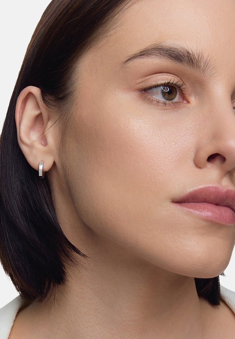 Srebrna kolczyk w kształcie kółka na gładkiej, jasnej karnacji; krótkie ciemne włosy otaczają twarz, podkreślając prosty design kolczyka i jego metaliczny blask.