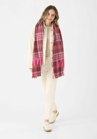 Rode en roze geruite sjaal met franjes aan de randen, voorzien van geborduurde tekst. Model draagt een lichtbeige outfit en sneakers. Achtergrond is wit.