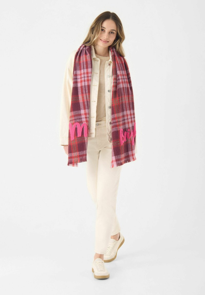 Rode en roze geruite sjaal met franjes aan de randen, voorzien van geborduurde tekst. Model draagt een lichtbeige outfit en sneakers. Achtergrond is wit.