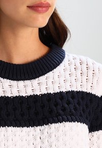 Garnittröja med marinblå ribbat krage och växelvis vita och marinblå texturerade ränder. Har ett vävt mönster för extra detalj.