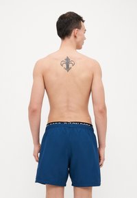 Navy blue swim shorts with an elastic waistband featuring a black logo. The wearer is shirtless, revealing a tattoo on the back.