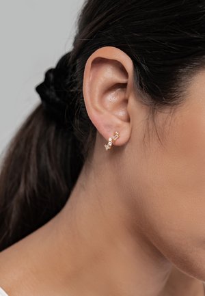 Goldfarbene Ohrstecker mit drei klaren Edelsteinen in unterschiedlichen Formen. Das Design zeichnet sich durch eine minimalistische, elegante Ästhetik aus.