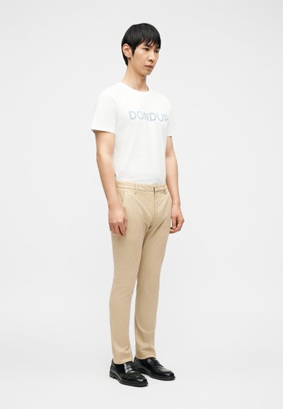 Άνδρας που φοράει λευκό t-shirt με την επιγραφή "DONDUP", μπεζ παντελόνι και μαύρα παπούτσια, στέκεται μπροστά σε απλό φωτεινό φόντο, κοιτάζοντας ελαφρώς προς τα δεξιά.