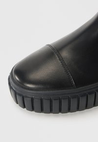 Bottine en cuir noir avec un bout rond, des coutures décoratives et une épaisse semelle en caoutchouc noir avec des rainures verticales.