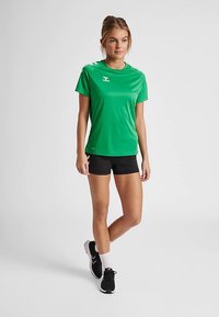 Groen atletisch T-shirt met korte mouwen, gladde textuur, ronde halslijn en logo; gecombineerd met zwarte shorts en zwarte hardloopschoenen.