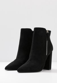 Stivali alla caviglia in suede nero con punta affilata, tacco spesso a blocco e dettaglio con zip laterale. Texture liscia con linee pulite e design semplice.