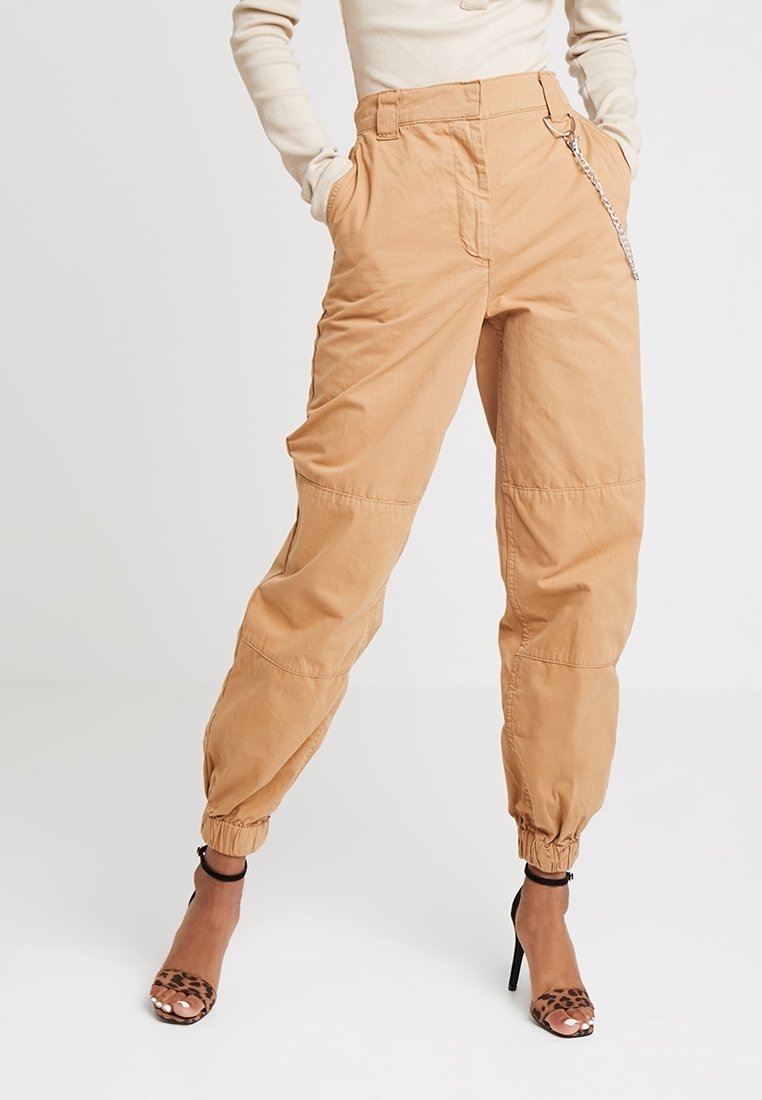 Pantalons cargo beiges avec un poignet élastique, des passants de ceinture et un détail en chaînage sur le côté ; fabriqués dans un tissu léger avec une coupe décontractée.