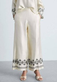 Satin wide-leg trousers in cream with black embroidered geometric patterns at the hem and pockets on each side. Smooth, flowy texture.