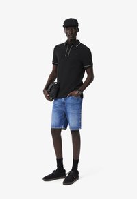 Cămașă polo neagră cu accente albe, pantaloni scurți din denim albastru, pantofi negri și o șapcă neagră, ținând o geantă mică neagră, stând pe un fond neutru.