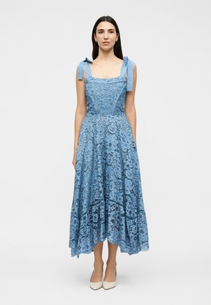 Kvinde iført en ærmeløs blå blonde midi-kjole med bundne skulderstropper, der står oprejst mod en ensfarvet lys baggrund.