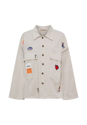 Beige skjortejakke med store forlommer, med mærker af et hjerte, goggles, nummer 7 og små tekstetiketter på ærmer og lommer.