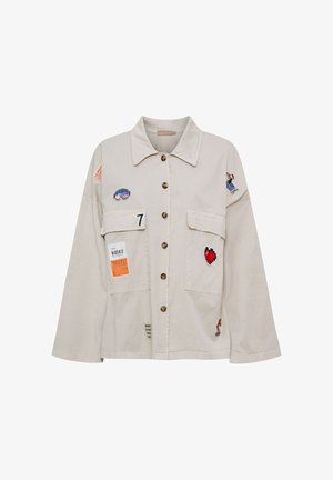 Beige skjortejakke med store forlommer, med mærker af et hjerte, goggles, nummer 7 og små tekstetiketter på ærmer og lommer.