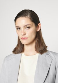 Blazer cinza claro sobre uma blusa de cor creme, com um design simples. Cabelo liso e de comprimento médio. Fundo neutro.