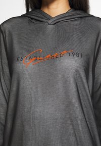 Sudadera negra con tejido texturizado, presenta el logo naranja de "Guess" con el texto "Established 1981" arriba en negro, diseñada con capucha ajustable con cordón.