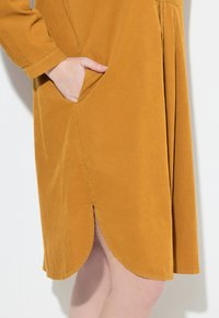 Senfgelbes Cordkleid mit abgerundetem Saum, Seitentasche und subtilen Falten details. Weicher Stoff mit lockerer Passform.