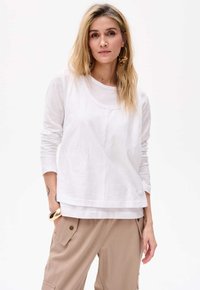 Weißes Langarmoberteil mit Rundhalsausschnitt, geschichteter Optik, glatter Textur und lockerer Passform, kombiniert mit beigen Cargo-Hosen.
