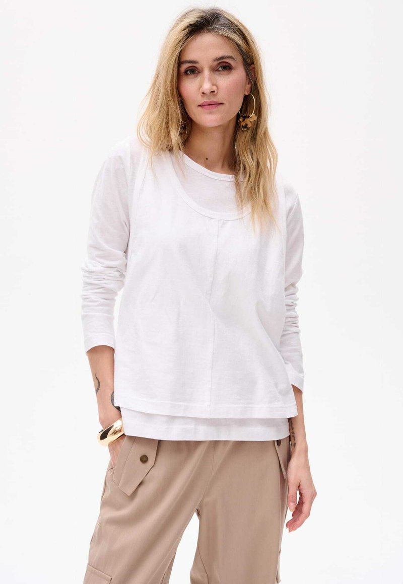 Weißes Langarmoberteil mit Rundhalsausschnitt, geschichteter Optik, glatter Textur und lockerer Passform, kombiniert mit beigen Cargo-Hosen.