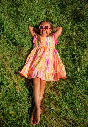 Buntes Kleid mit geometrischen Mustern in Pink, Orange und Grün; runde rosa Sonnenbrille; Kind, das auf grünem Gras liegt und lächelt.