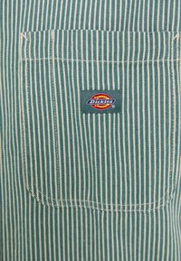 Blue-striped work shirt featuring a square chest pocket and a sewn-on logo patch with a circular design in red, yellow, and blue.