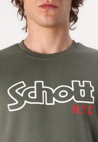 Maglietta girocollo verde oliva con grande logo "Schott N.Y.C." bianco e rosso in carattere audace. Tessuto di cotone liscio con una forma aderente.