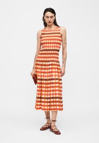 Femeie cu tatuaje, purtând o rochie fără mâneci în carouri portocalii, ținând o poșetă maro și purtând pantofi maro cu șireturi, stând în fața unui fundal alb.