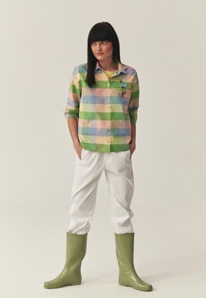 Žena s čiernymi vlasmi v pastelovej kockovanej košeli, bielych nohaviciach a svetlozelených gumákoch stojaca s rukami v vreckách.